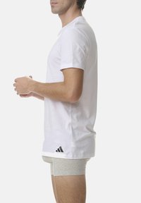 White cotton t-shirt with short sleeves and a round neckline, featuring a black Adidas logo near the hem. Gray underwear is visible underneath.