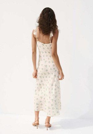 Kobieta o kręconych włosach nosi kremową sukienkę midi w kwiaty z falbanianymi ramiączkami, stoi boso na jednolitym białym tle.