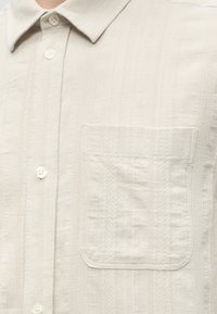 Chemise beige texturée à boutons avec col pointu et poche carrée sur la poitrine côté gauche.