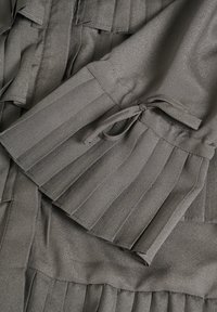 Tessuto plissettato grigio con una morbida lucentezza, con maniche gather legate al polso e dettagli strutturati. Design elegante e testurizzato.
