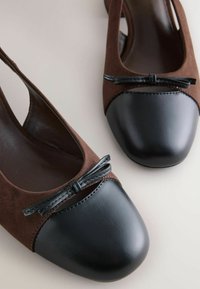 Brūni zamšādas slingback apavi ar melnu ādas purngali, priekšpusē ar mazu melnu loku akcentu. Gluda tekstūra un noapaļota forma.