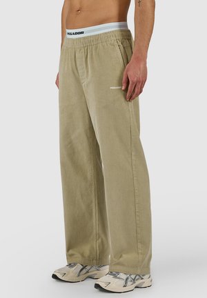 Mężczyzna ubrany w beżowe szerokie spodnie sztruksowe z elastycznym pasem i białoszare sneakersy, stojący na jednolitym tle.