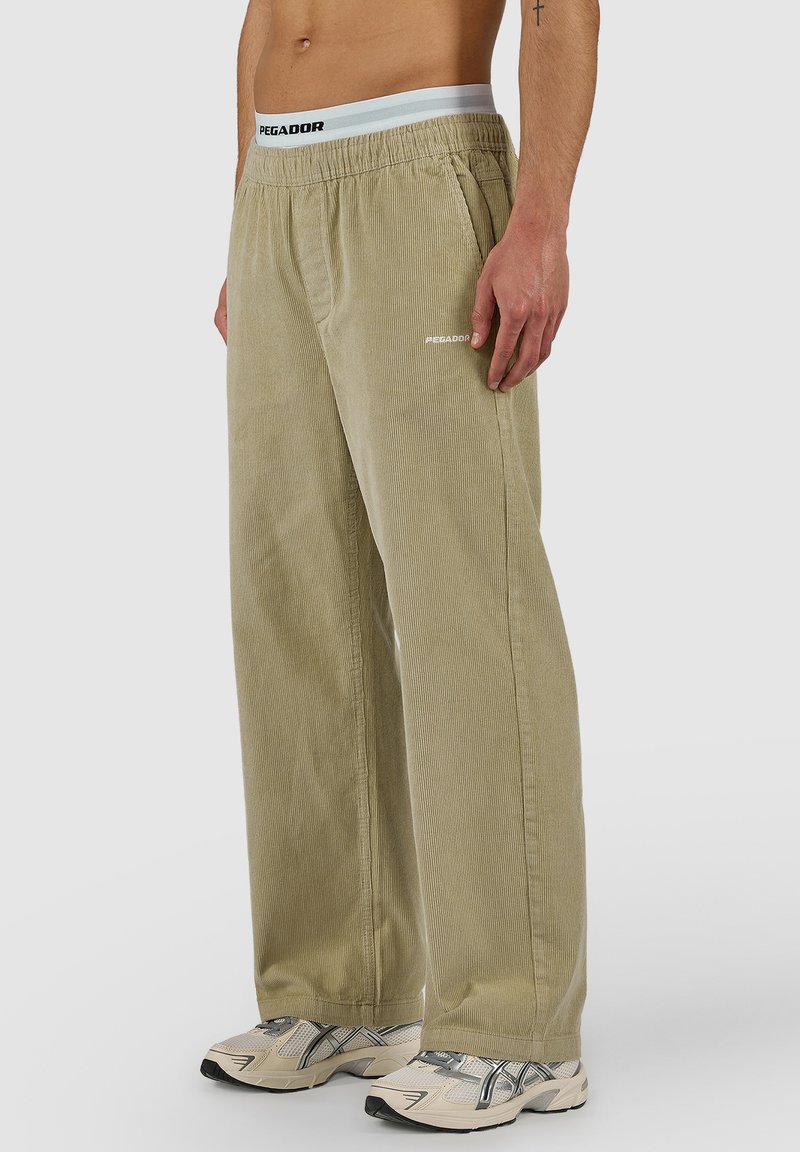 Hombre con pantalones anchos de pana beige con cintura elástica y zapatillas blancas-grises, de pie frente a un fondo liso.