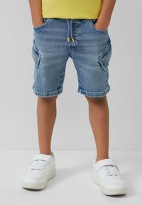 Pantalones cortos de mezclilla en azul claro con cintura ajustable por cordón, bolsillos frontales y bolsillos laterales tipo cargo. Llevados con zapatillas blancas, mostrando un estilo casual.
