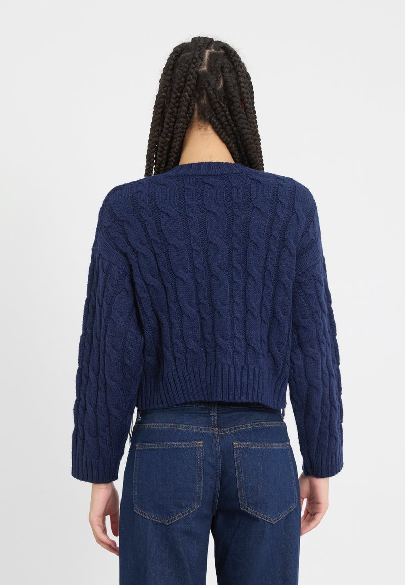 Pull à mailles torsadées bleu marine au design court, avec un col rond et des poignets côtelés, associé à un jean en denim foncé.