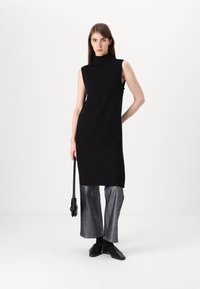 Μαύρο χωρίς μανίκια φόρεμα με ψηλό λαιμό, λιτή σχεδίαση, μήκους μέχρι το �γόνατο, συνδυασμένο με μεταλλικά παντελόνια με φαρδιά πόδια και μαύρα παπούτσια χωρίς λουράκι.