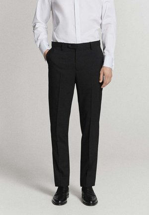 Homme portant un pantalon noir sur mesure, une chemise blanche et des chaussures en cuir noires, debout avec une main dans la poche.