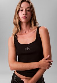 Μαύρο ριμπ αμάνικο τοπ με τετράγωνο λαιμόκοψη και λεπτομέρεια λογότυπου. Το ύφασμα έχει λεία υφή, παρουσιάζεται σε μοντέλο με σταυρωμένα χέρια.