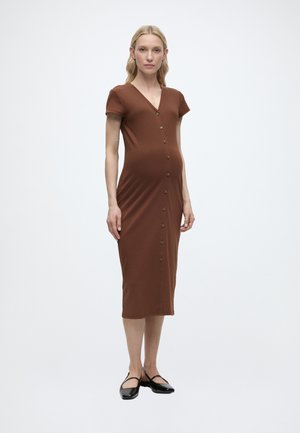 Tehotná žena stojaca, oblečená v priliehavých hnedých rebrovaných midi šatách s gombíkmi vpredu a čiernymi plochými topánkami.