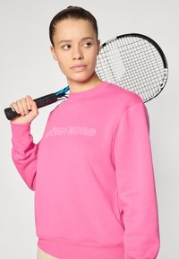 Rosa hettegenser med rund hals, med "BJÖRN BORG" logo i hvitt. Modellen holder et tennisslag, og viser en avslappet sportstil.