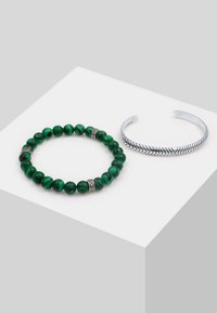 Bracciale di perline in malachite verde con accentuazioni in argento accanto a un bracciale in argento con un motivo a spina di pesce.