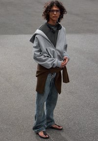 Grå oversized hoodie, mørk t-shirt, brun sweater bundet om livet, blå jeans, sorte flip-flops, iført høretelefoner og briller.