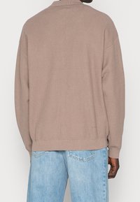 Uomo che indossa un maglione di lana beige e jeans di denim blu chiaro, visto da dietro su uno sfondo semplice.