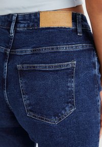 Mørkeblå denimjeans med lærmerke, baklomme-design og synlige søm detaljer. Glatt tekstur med en tettsittende form.