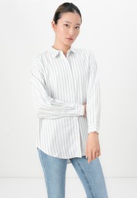 Hvit langermet skjorte med svarte vertikale striper, knappelukking foran, krage og lett buet kant; sammen med lyseblå jeans.
