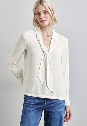 Vit blus med tie-krage, långa ärmar och en slät textur. Matchas med blå jeans, vilket visar en avslappnad och mångsidig stil.