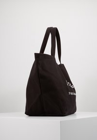 Black canvas tote bag with two long handles, a spacious body, and white text detailing on one side. Smooth texture, minimalistic design.