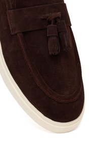 Donkerbruine suède loafer met kwastjes en een witte rubberen zool, met gedetailleerde stiksels op de teen en het bovenste voorste gedeelte.