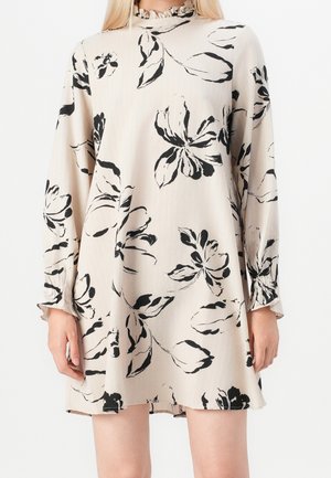 Robe beige à manches longues avec imprimé floral noir, col haut volanté et poignets froncés, portée au-dessus des genoux.