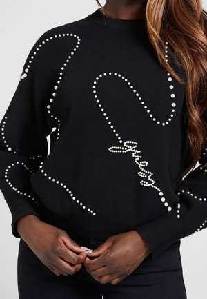 Pull noir avec des embellissements en forme de perles formant des arabesques abstraites et le mot « Guess », porté par une personne aux longs cheveux ondulés.
