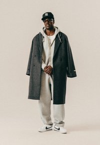 Homme portant une casquette noire, des lunettes, un long manteau sombre par-dessus un sweat à capuche gris et un pantalon de jogging, des baskets blanches, se tenant contre un fond clair uni.