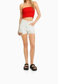 Top rojo sin tirantes con volantes texturizados y shorts de mezclilla blanca con bordes deshilachados. Sandalias de plataforma negras con hebillas gruesas.