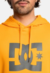 Žltá mikina s kapucňou a šedým logom DC, so šnúrkami a kovovými akcentmi. Mäkkučký materiál s voľným strihom.