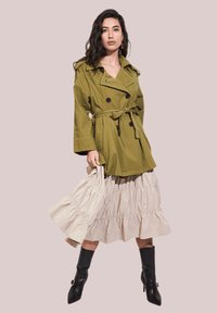 Trench coat color oliva con cinturón y botones de doble botonadura, falda beis con volantes en capas, y botas negras hasta la rodilla. Texturas de tela suaves.