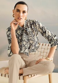 Frau mit nach hinten gegeltem Haar trägt eine Bluse mit Schlangenmuster, beige Hosen, goldene Creolen und ein Armband und sitzt im Schneidersitz auf einem geflochtenen Stuhl.