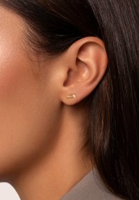Boucles d'oreilles en forme d'os de poisson doré avec un design texturé et une petite pierre d'accent, portées sur l'oreille d'un modèle au teint lisse.