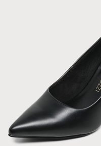 Γυναικείο μαύρο παπούτσι με ψηλό τακούνι και μυτερή μύτη, με λείο δερμάτινο επάνω μέρος και μαλακό ένθετο πάτο σε ουδέτερο φόντο.