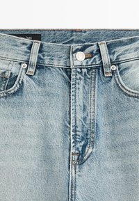 Jean en denim bleu clair au niveau de la taille avec passants de ceinture, bouton en métal, fermeture éclair, poches avant et détails de coutures visibles.