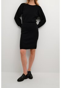 Pull en maille noir avec de longues manches bouffantes associé à une jupe noire cintrée jusqu'au genou. Porté avec des chaussures noires à enfiler.