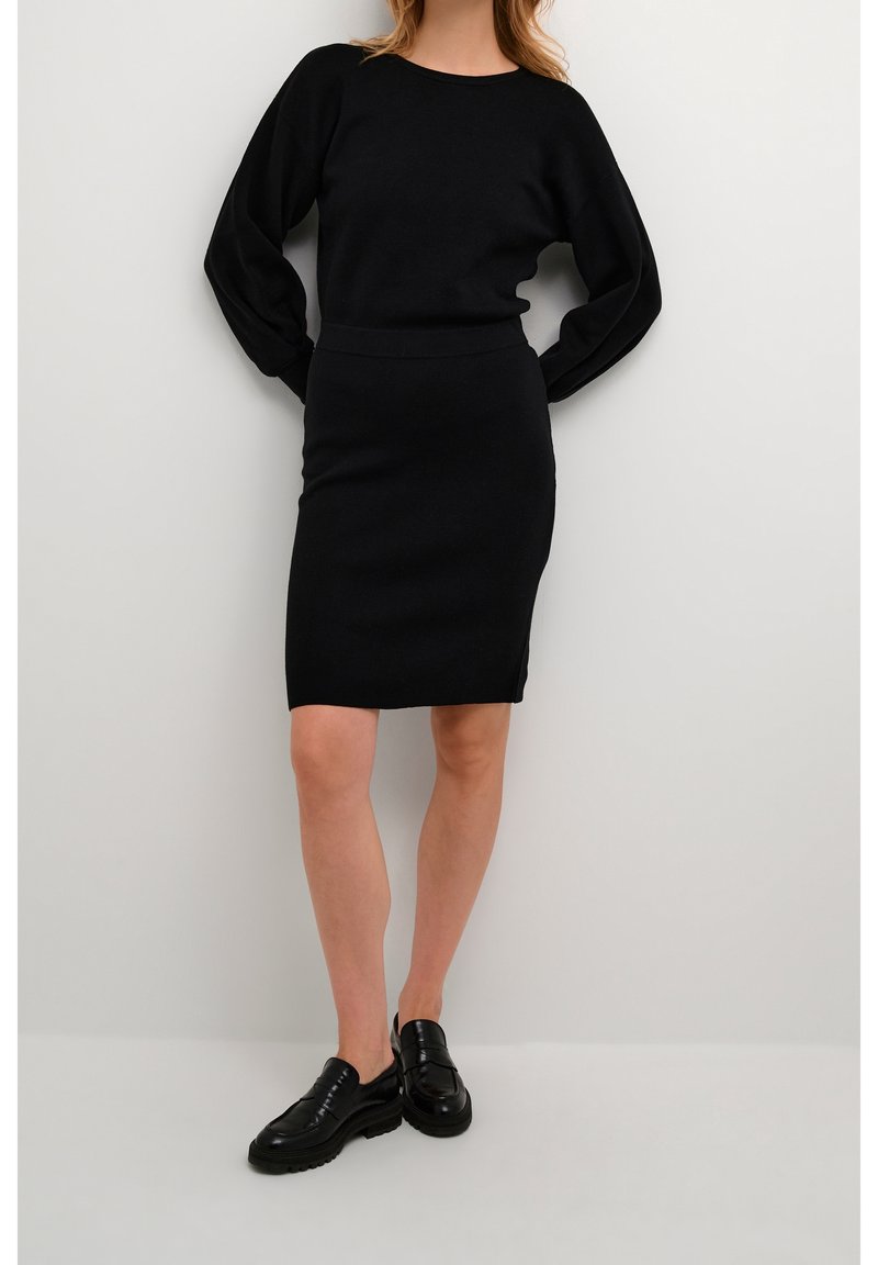 Pull en maille noir avec de longues manches bouffantes associé à une jupe noire cintrée jusqu'au genou. Porté avec des chaussures noires à enfiler.