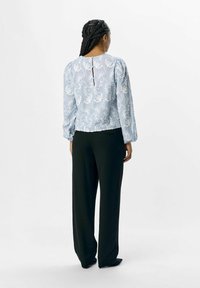 Blusa de color azul claro con bordado floral blanco, mangas acampanadas y detalle de ojal en la parte posterior, combinada con pantalones negros de talle alto.