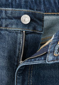 Gros plan d'un jean en denim bleu montrant une fermeture éclair partiellement ouverte et un bouton métallique avec des détails de couture.