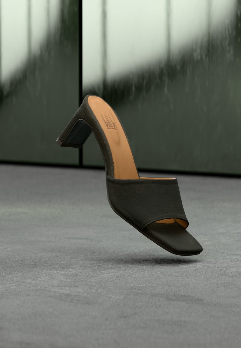 Mule noire à bout ouvert avec un bout carré et un talon moyen à bloc, posée sur un tapis gris avec des panneaux verts flous en arrière-plan.