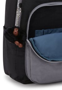 Schwarzer Stoff-Rucksack mit einer grauen Reißverschluss-Vortasche und einem blauen Innenraum. Mit einem flauschigen schwarzen Affen-Schlüsselanhänger, der am Reißverschluss befestigt ist.