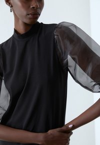 Top negru cu guler înalt și mâneci puff transparente. Corpul este realizat dintr-un material neted, în timp ce mânecile au o textură moale și translucidă.