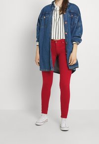 Giacca camicia in denim, azzurro chiaro con bottoni di rame, indossata sopra un top corto a righe bianche. Jeans rossi attillati e sneakers bianche completano l'outfit.