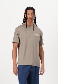 Kortermet t-skjorte i tan laget av glatt stoff, med en liten trykt logo på brystet, kombinert med blå denimjeans.