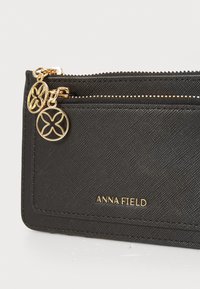 Portefeuille en tissu noir avec fermeture éclair, accents en matériel doré et logo embossé "ANNA FIELD" à l'avant. Fini texturé.