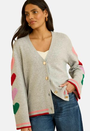Mladá žena s dlhými tmavými vlasmi, oblečená v sivom zapínacom kardigane ozdobenom farebnými srdiečkami na rukávoch a modrých džínsoch.