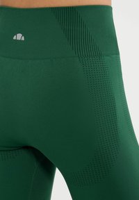 Los leggings verdes presentan un material suave con acentos texturizados, incluyendo paneles de malla y patrones de puntos a lo largo de la cinturilla y los lados.