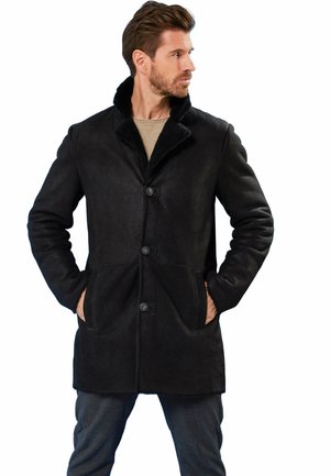 Mann trägt einen schwarzen Mantel mittlerer Länge mit Knopfleiste und Pelzkragen, Hände in den Taschen, schaut nach rechts vor weißem Hintergrund.