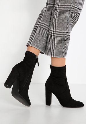 Black suede ankle boots with block heels and side zippers worn with cropped black-and-white plaid pants.