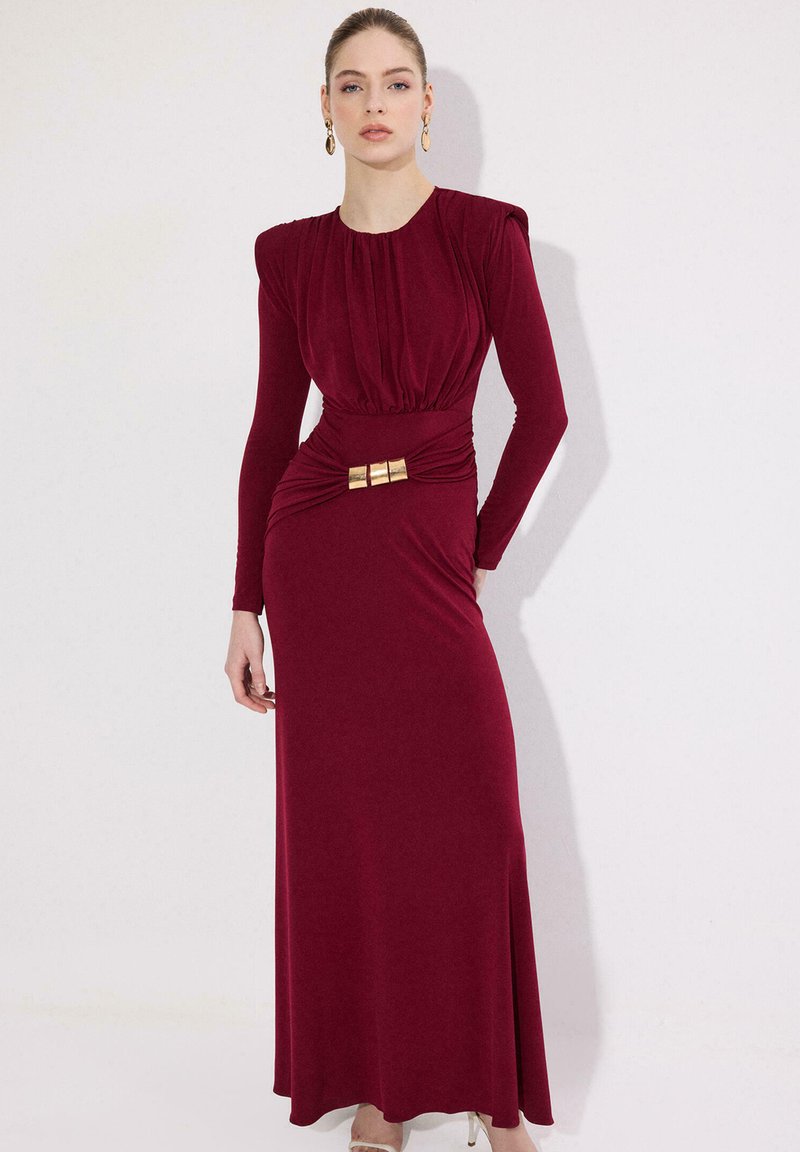 Γυναίκα που φοράει μακριά μανίκια, φόρεμα μήκους μέχρι το πάτωμα σε μπορντό χρώμα με πλισέ μπούστο και χρυσή αγκράφα στη μέση, στέκεται μπροστά σε λευκό τοίχο.