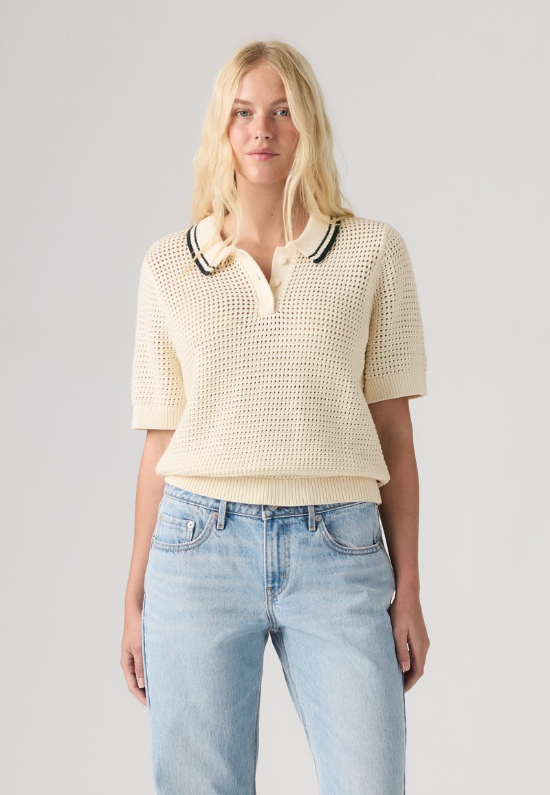 Femme blonde portant un polo en maille crème à manches courtes avec un col orné d'une bordure noire et un jean taille haute bleu clair, debout devant un fond uni.