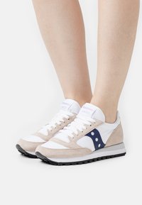 Saucony Jazz tenisice imaju bež antilop i bijelu kožnu gornjište, plave akcentne detalje i teksturiranu crnu gumenju podlogu.