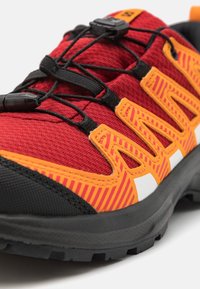 Scarpa sportiva rossa e arancione con una parte superiore in rete testurizzata, suola in gomma nera, lacci elastici e un evidente dettaglio a strisce.